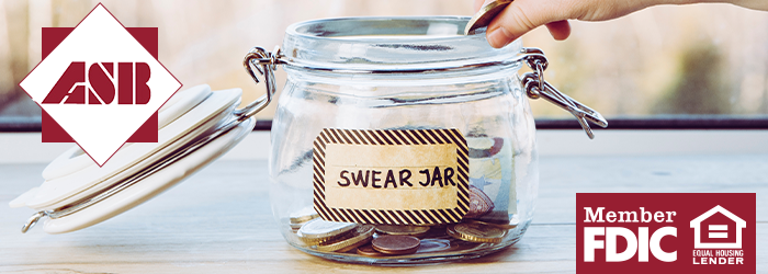 Glass jar with label 'Swear Jar' on it and coins in it with person's hand dropping another coin in