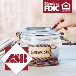 Glass jar with label 'Swear Jar' on it and coins in it with person's hand dropping another coin in